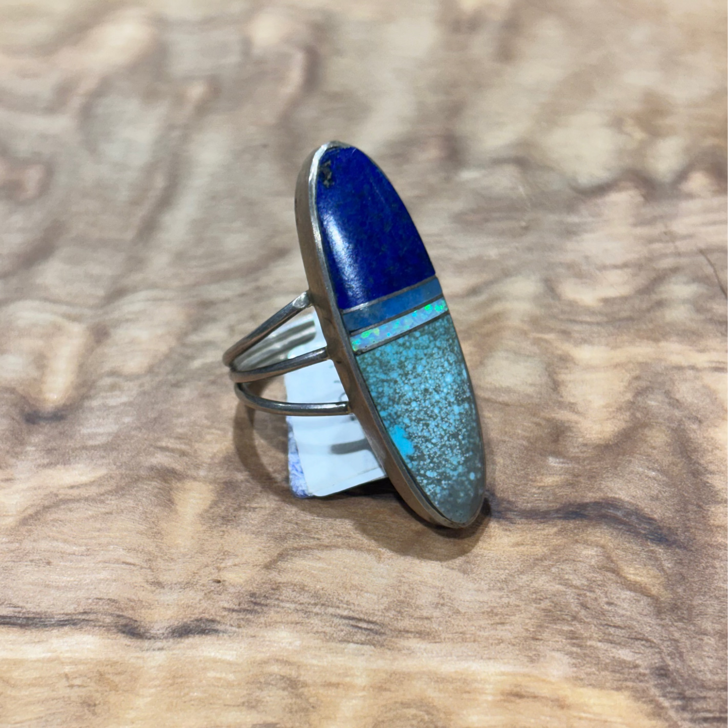 A silver ring with a large oval inlay featuring a deep blue top section and a turquoise-blue speckled bottom section, displayed on a textured wood surface.