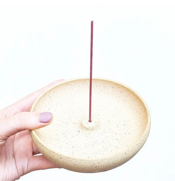 Hand holding a beige ceramic incense holder with a single incense stick on a white background