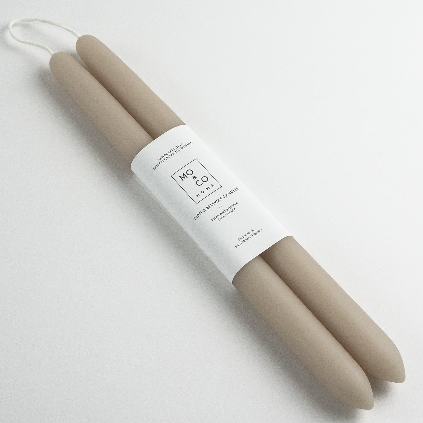 Two beige candles in a white cylindrical holder with 'MO&CO' branding on a light gray background.