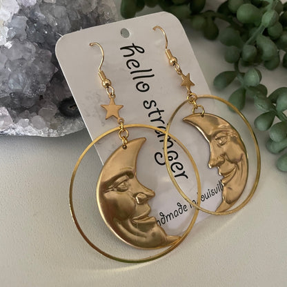 A pair of gold-toned statement earrings with large hoop rings and crescent moon faces, displayed on a white card with decorative crystals and greenery in the background.