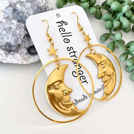 A pair of gold-toned statement earrings with large hoop rings and crescent moon faces, displayed on a white card with decorative crystals and greenery in the background.