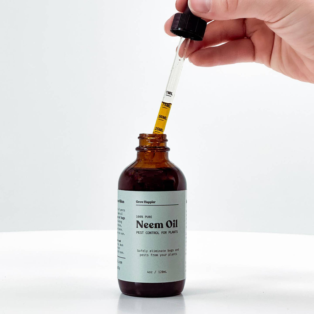 A hand holds a glass dropper above an amber glass bottle labeled Neem Oil, with the dropper containing yellow oil about to be dispensed into the bottle. The bottle has a white label and a black cap, set against a clean white background.