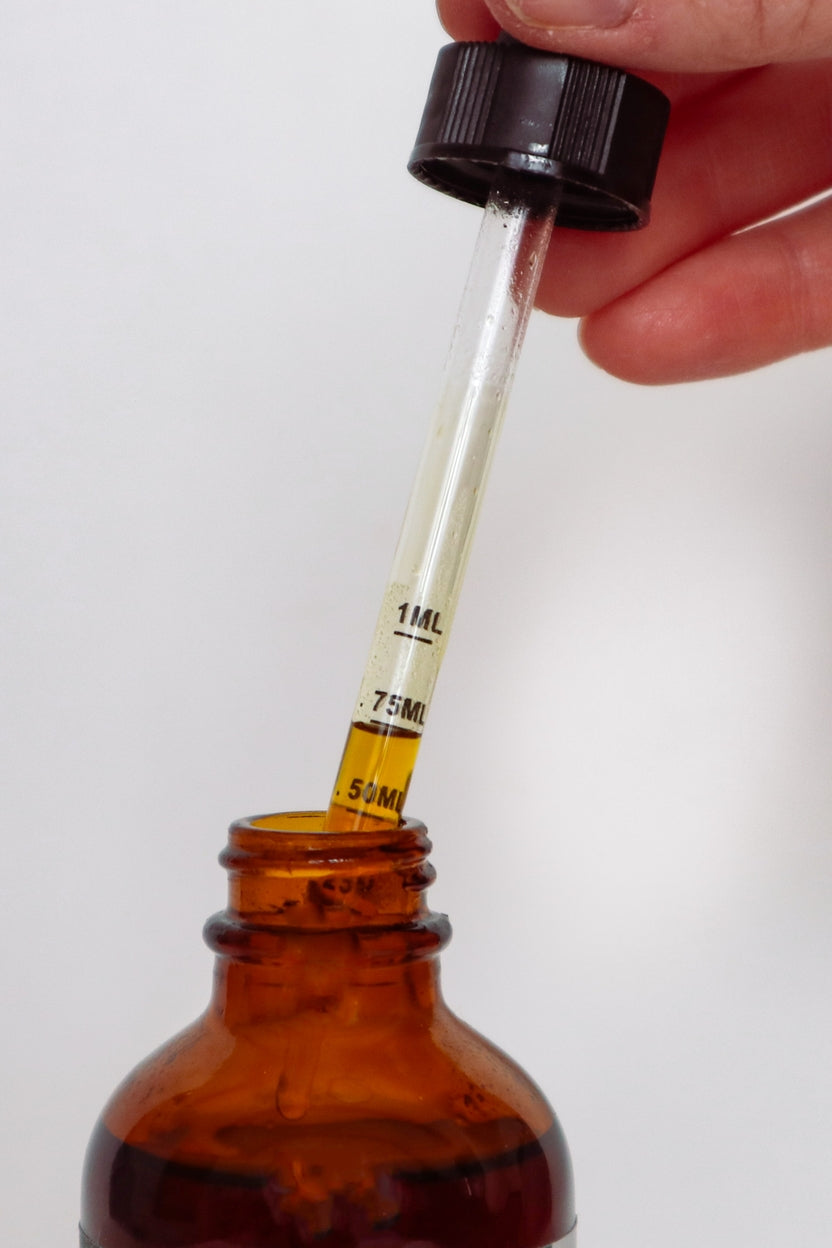 A close-up of a hand holding a glass dropper above an amber glass bottle, with liquid being transferred from the dropper to the bottle. The dropper has black measurement markings and a black cap, set against a plain light background.
