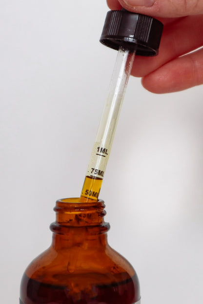 A close-up of a hand holding a glass dropper above an amber glass bottle, with liquid being transferred from the dropper to the bottle. The dropper has black measurement markings and a black cap, set against a plain light background.