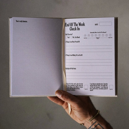 Hand holding an open book with a 'End of the Week Check In' form on a gray background