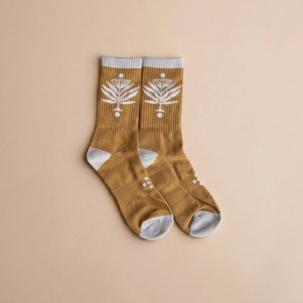 A pair of mustard brown ribbed crew socks with white knit botanical patterns on the upper leg, white heels, toes, and cuffs, displayed on a solid beige background.