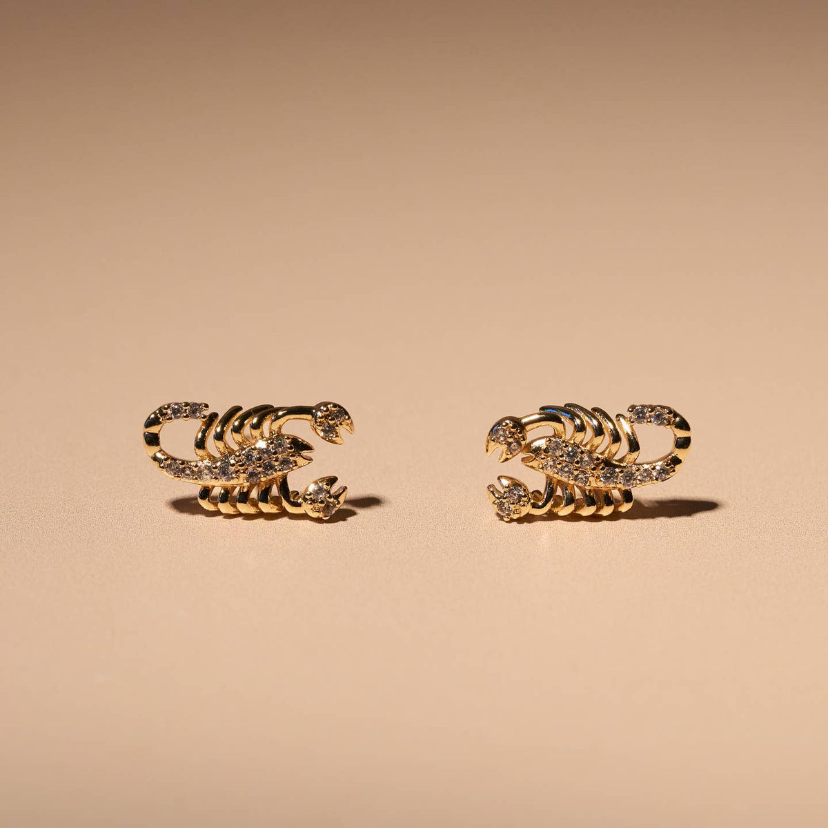 A pair of gold-toned scorpion stud earrings embellished with small clear crystals, displayed on a warm beige background.