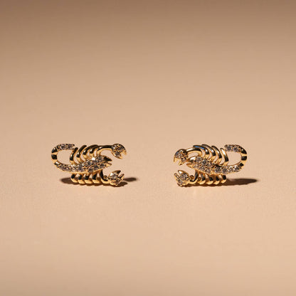 A pair of gold-toned scorpion stud earrings embellished with small clear crystals, displayed on a warm beige background.