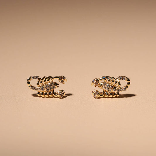 A pair of gold-toned scorpion stud earrings embellished with small clear crystals, displayed on a warm beige background.