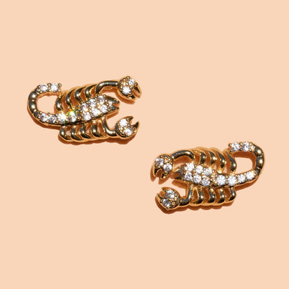A pair of gold-toned scorpion stud earrings embellished with clear pavé gemstones, set against a solid pale peach background.