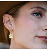 A close-up of a woman's ear wearing a delicate gold hoop earring with a small round gold disc charm.