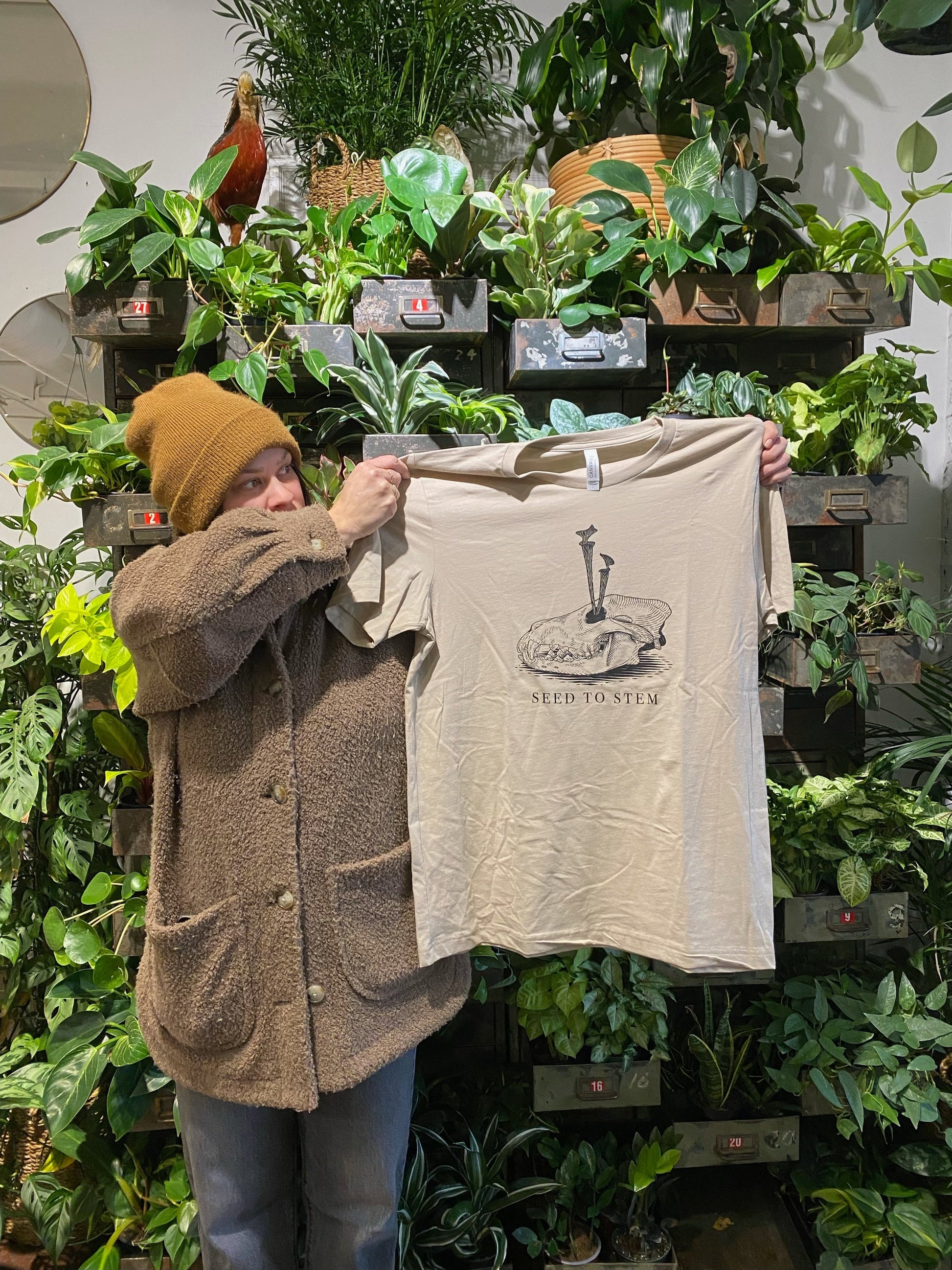 Person holding a beige t-shirt with a design in front of a plant wall.