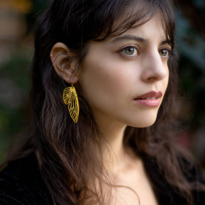 A close-up portrait of a woman wearing a single gold leaf-shaped dangle earring with an intricate openwork design, set against a soft, blurred green background.