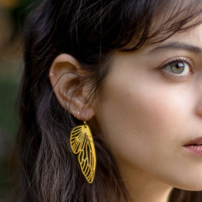 A close-up of a woman's ear wearing a delicate gold wire earring shaped like a wing, with her dark hair and green eyes visible against a softly blurred outdoor background.