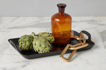 Two artichokes on a black tray with an amber bottle and gold tool on a marble surface