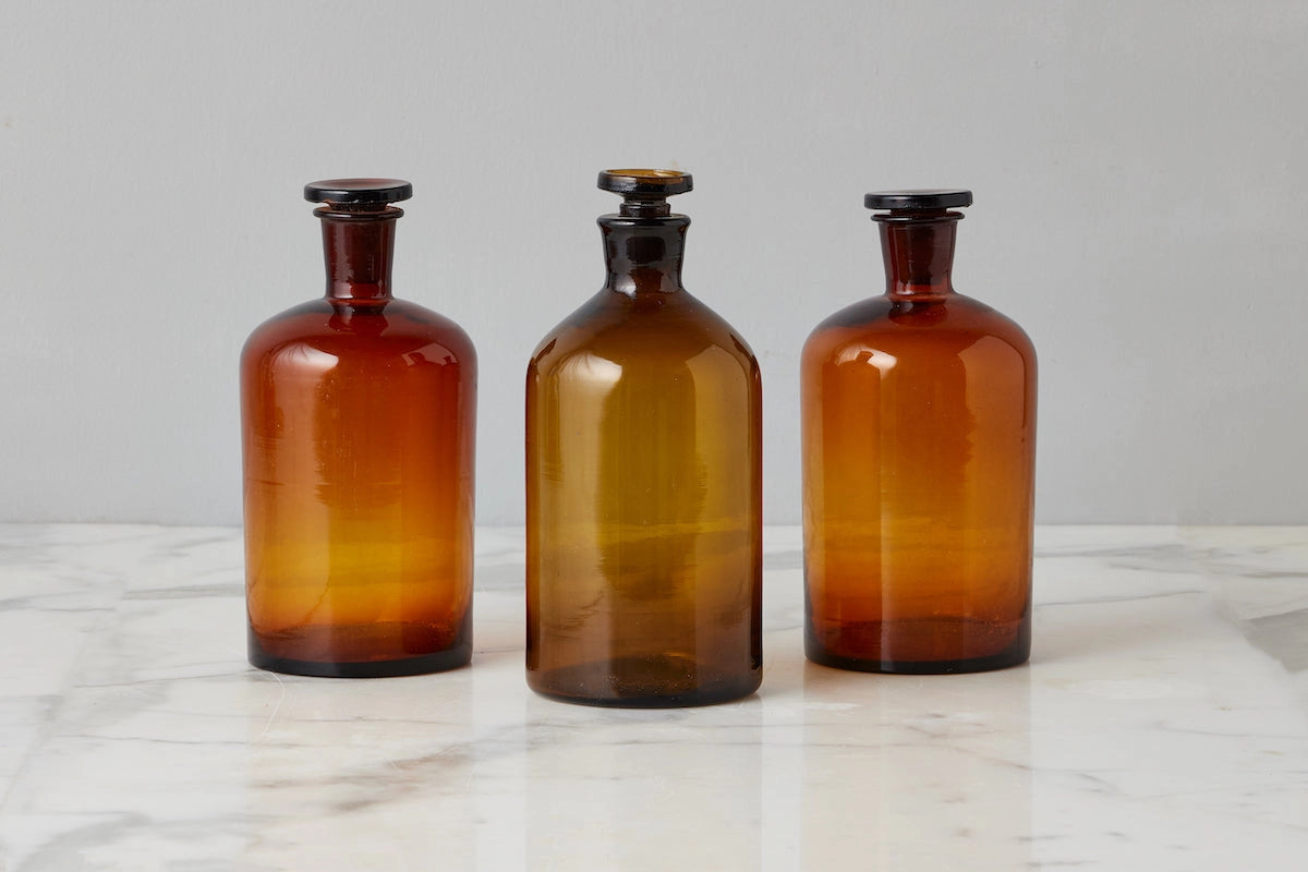 Three amber glass apothecary-style bottles with dark stoppers, arranged side by side on a light marble surface against a neutral gray background.