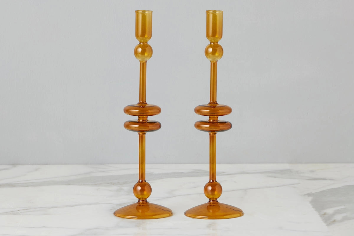 A pair of amber glass candlesticks with a mid-century modern design, featuring stacked circular rings and a flared base, set against a neutral gray background and white marble surface.