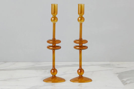 A pair of amber glass candlesticks with a mid-century modern design, featuring stacked circular rings and a flared base, set against a neutral gray background and white marble surface.