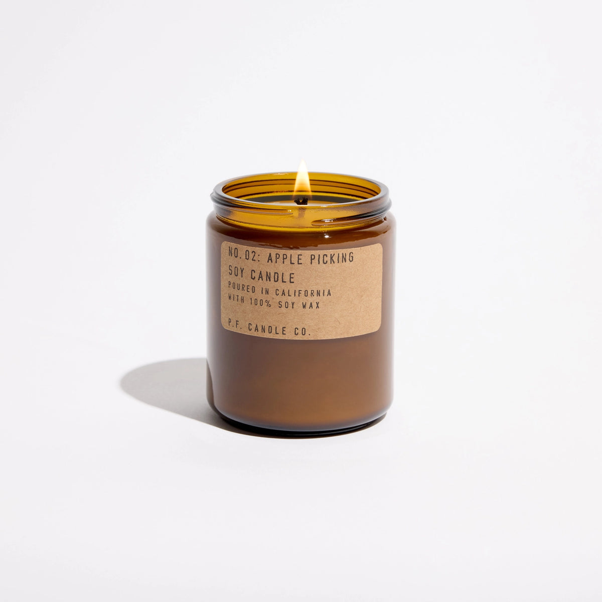 An amber glass jar candle with a lit wick, featuring a kraft paper label that reads '16 OZ. APPLE PICKING SOY CANDLE'.