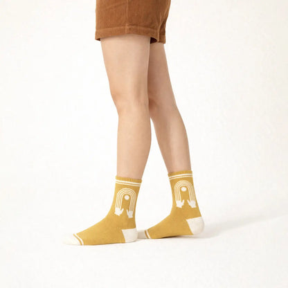 A person wearing mustard yellow socks with a white arch graphic and white toe/heel sections, paired with brown shorts, shown against a plain white studio background.