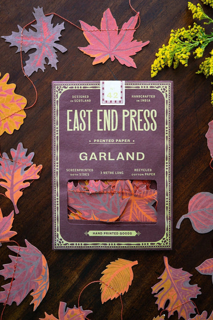 A package of East End Press Garland printed paper sheets lies on a dark wooden surface surrounded by colorful autumn leaves and small yellow flowers. The package is mauve-purple with cream text and border details, and a window near the bottom shows folded paper samples in pink, orange, and red tones.