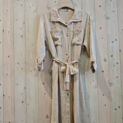 A light beige linen shirt dress with a collar, button front, chest pockets, and a self-tie belt, hanging on a wooden hanger against a light wood plank wall.