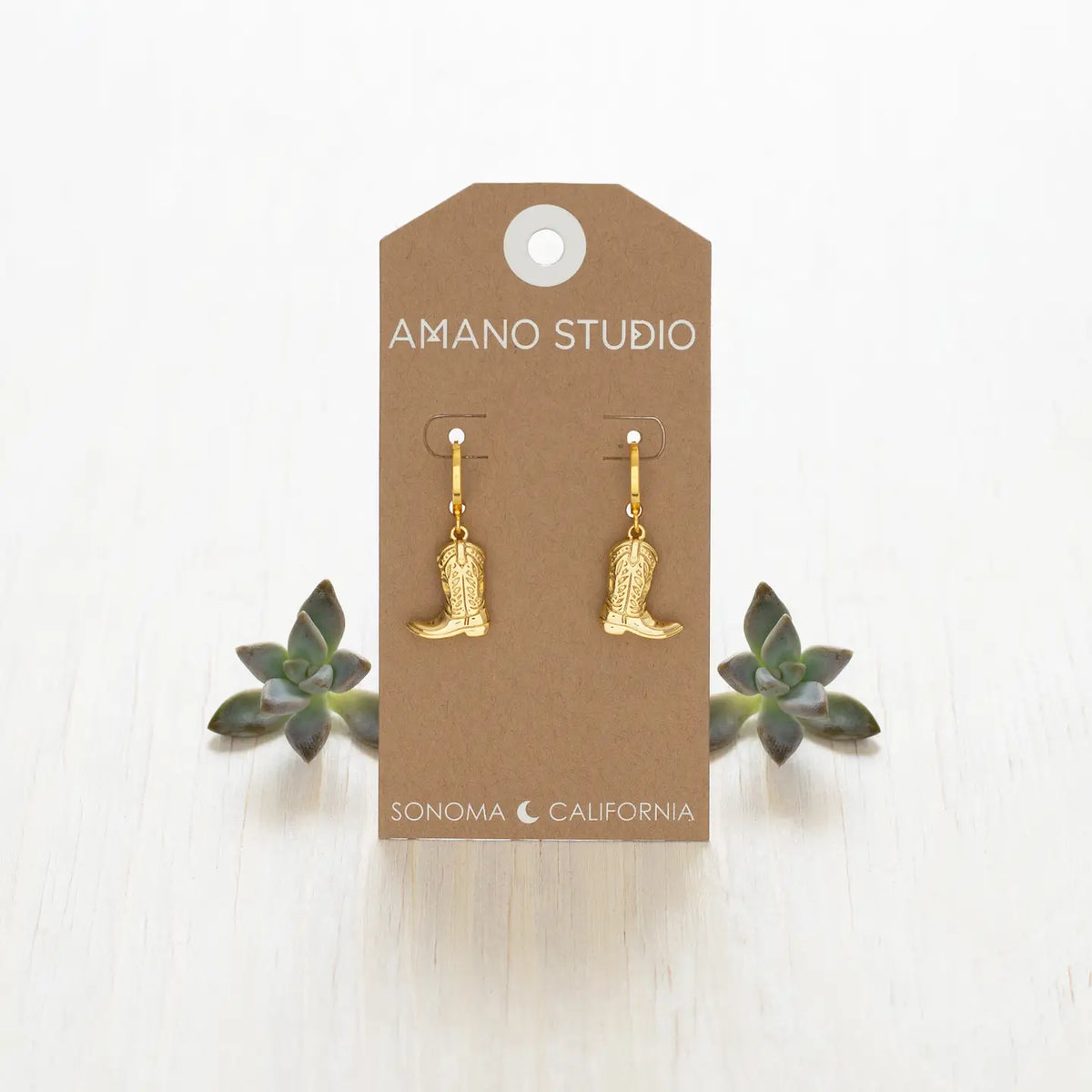 Gold cowboy boot earrings on a 'Amano Studio' branded card with succulents in the background.
