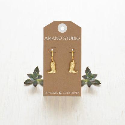 Gold cowboy boot earrings on a 'Amano Studio' branded card with succulents in the background.