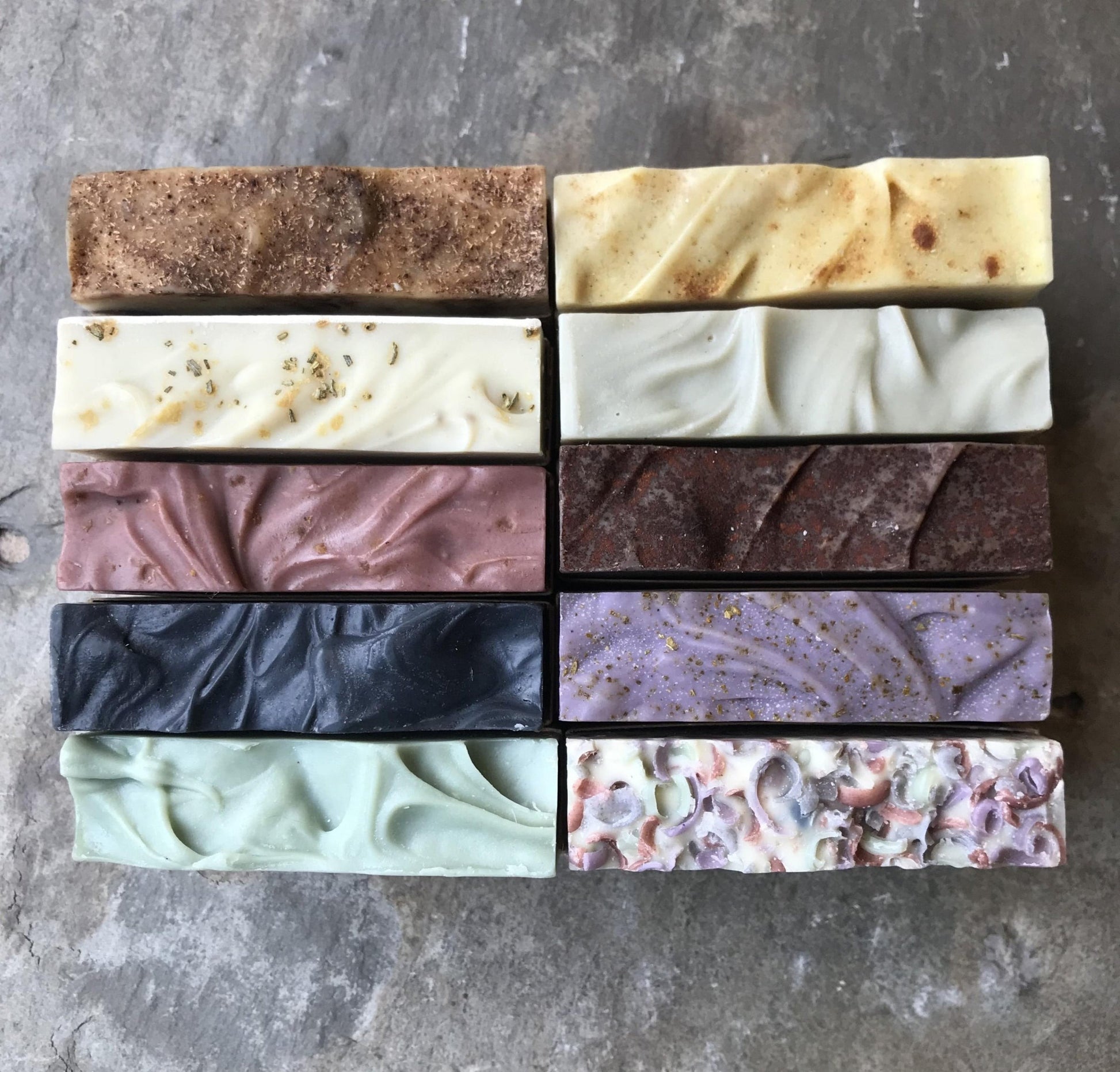 An array of natural handmade soap bars with varying colors and textures, displayed in four rows on a textured grey surface.
