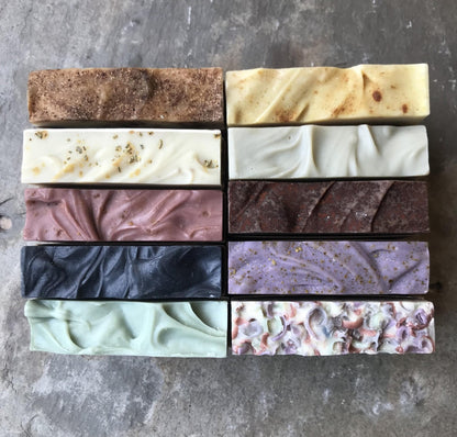 An array of natural handmade soap bars with varying colors and textures, displayed in four rows on a textured grey surface.