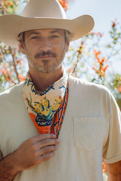 Man wearing a wide-brimmed hat and colorful bandana, standing outdoors with trees in the background.