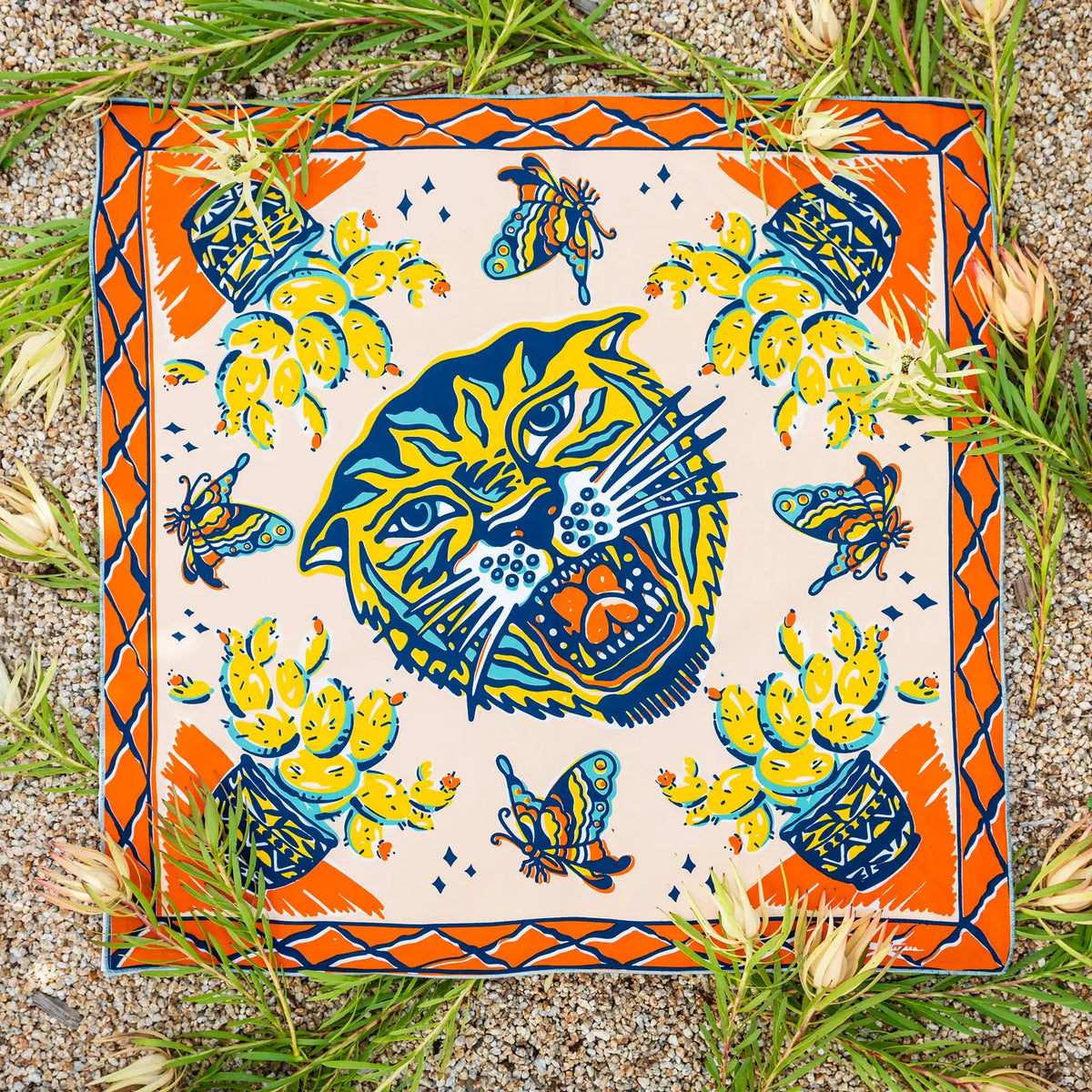 A square ceramic tile or coaster featuring a colorful Mexican-style illustration of a roaring jaguar head, surrounded by butterflies, cacti, and a patterned border, photographed flat on a gravel surface with green grass and small flowers around it.
