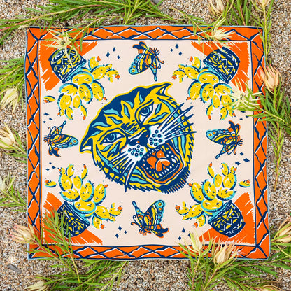 A square ceramic tile or coaster featuring a colorful Mexican-style illustration of a roaring jaguar head, surrounded by butterflies, cacti, and a patterned border, photographed flat on a gravel surface with green grass and small flowers around it.