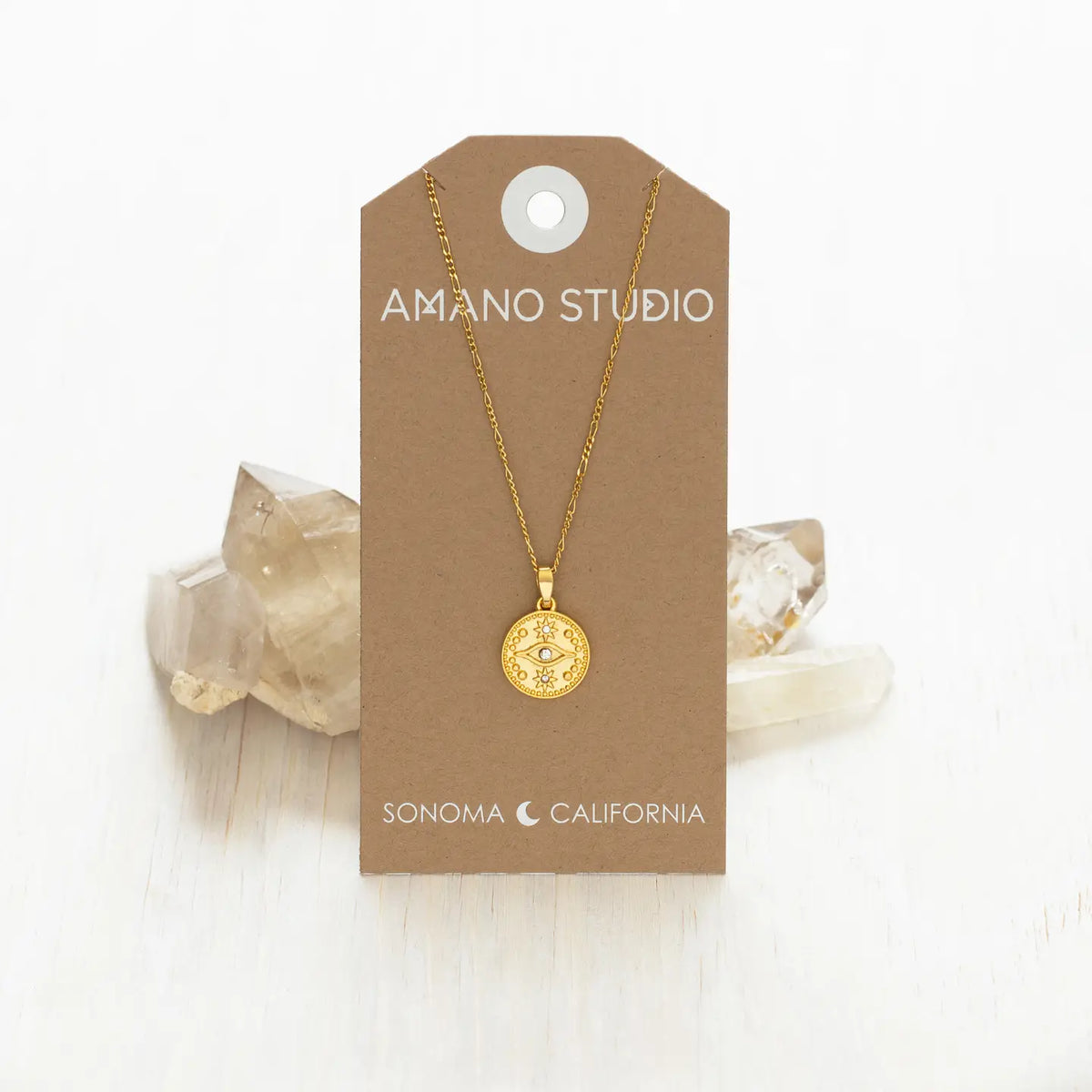 A gold pendant necklace displayed on a brown kraft card labeled 'AMANO STUDIO' and 'SONOMA CALIFORNIA,' photographed on a light wood surface with clear crystal clusters behind it.