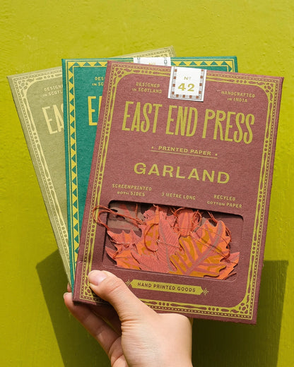 A hand holds three East End Press printed paper garland boxes in green and brown tones against a bright yellow-green background. The front box is maroon with gold lettering and shows orange-red paper leaf garland inside a window.