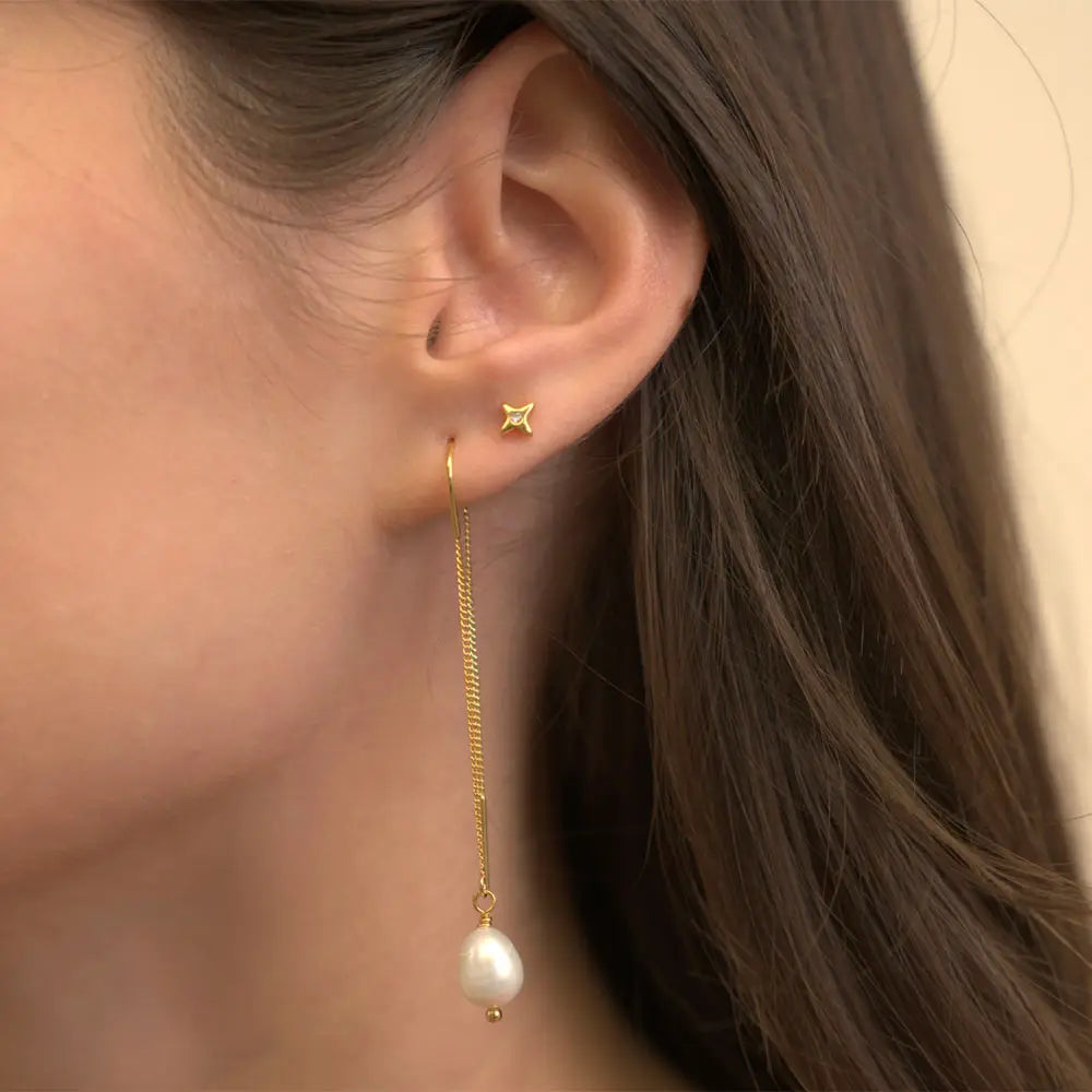 A close-up of a model's ear wearing a delicate gold dangle earring with a small star-shaped stud and a long chain ending in a white pearl, against a soft beige background.