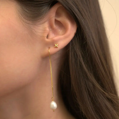 A close-up of a model's ear wearing a delicate gold dangle earring with a small star-shaped stud and a long chain ending in a white pearl, against a soft beige background.