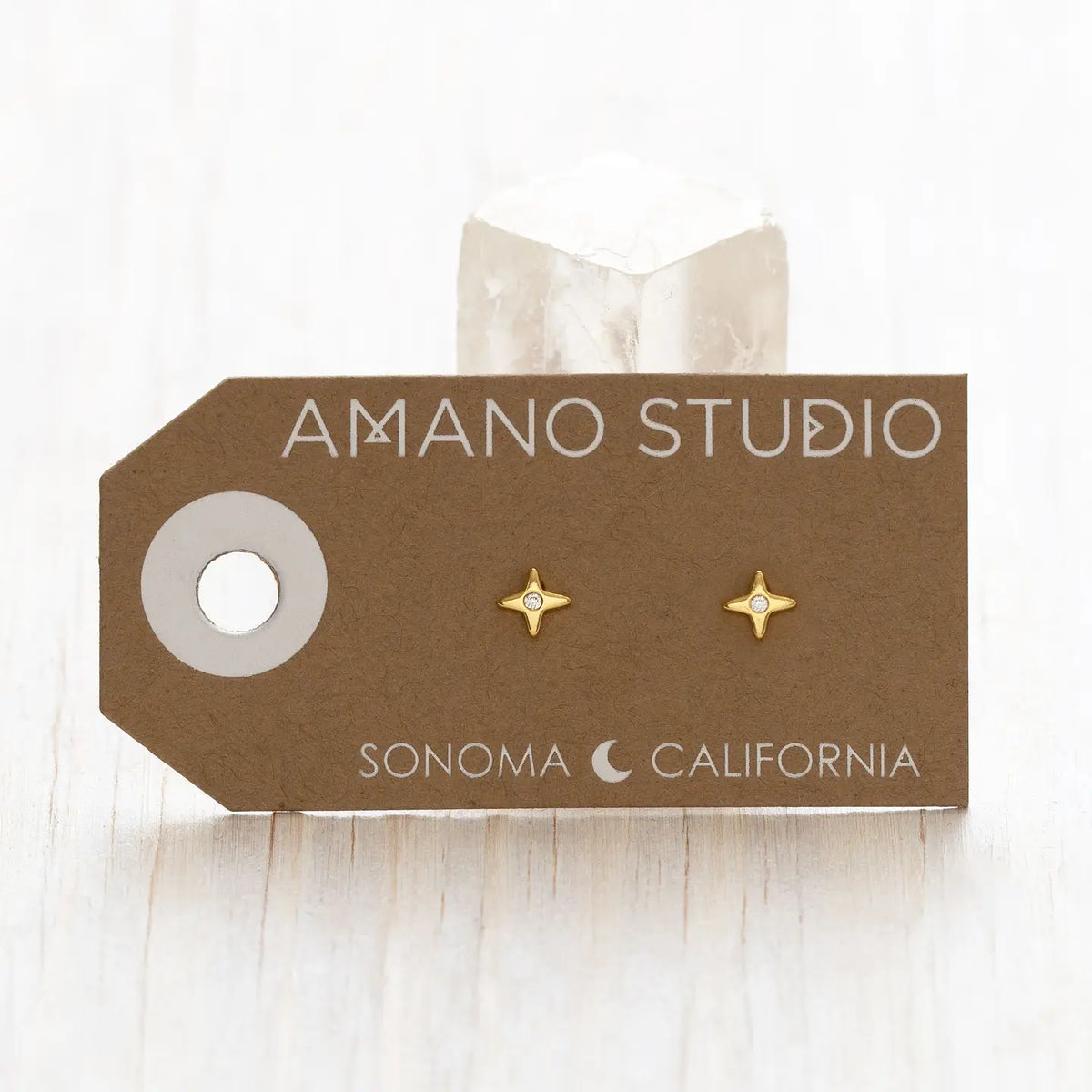 A pair of small gold starburst stud earrings displayed on a brown kraft card labeled AMANO STUDIO and SONOMA CALIFORNIA, with a clear crystal behind the card on a light wood surface.