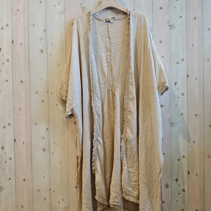 A light beige, crinkled cotton-linen open-front cardigan or kaftan hanging on a wooden hanger against a light wood plank wall.