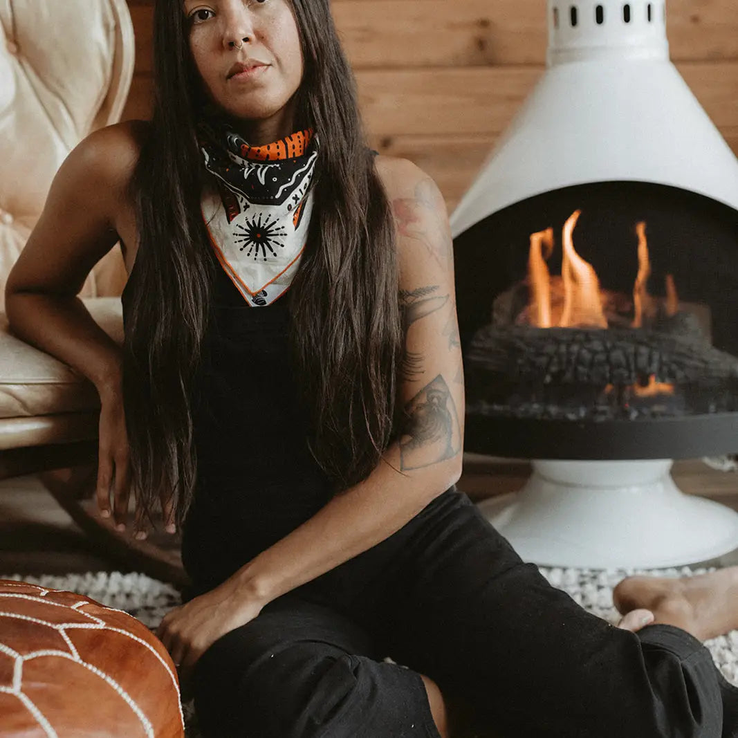 A woman with long dark hair sits on a white stone fireplace hearth in a rustic room with wood-paneled walls. She is wearing a black sleeveless top and black pants, with a white bandana featuring orange and black graphics wrapped around her neck.