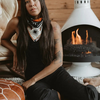 A woman with long dark hair sits on a white stone fireplace hearth in a rustic room with wood-paneled walls. She is wearing a black sleeveless top and black pants, with a white bandana featuring orange and black graphics wrapped around her neck.