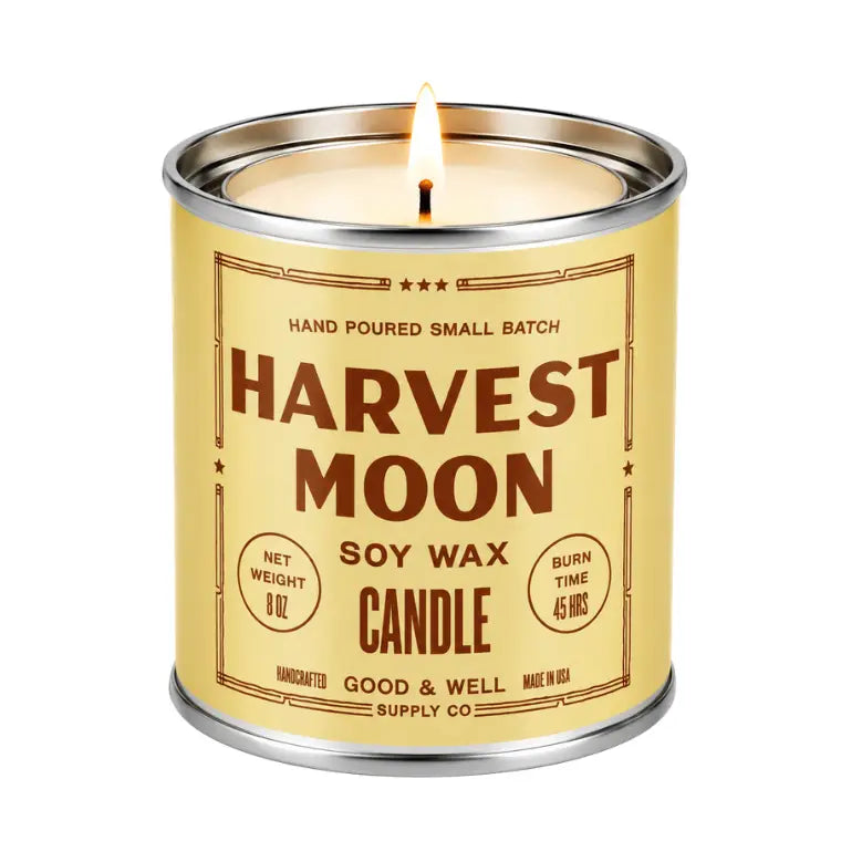 A lit soy wax candle in a cylindrical metal tin with a cream-colored label and silver metal rim, shown on a plain white background. The label reads 'HARVEST MOON' and 'SOY WAX CANDLE' with 'GOOD & WELL SUPPLY CO' at the bottom.