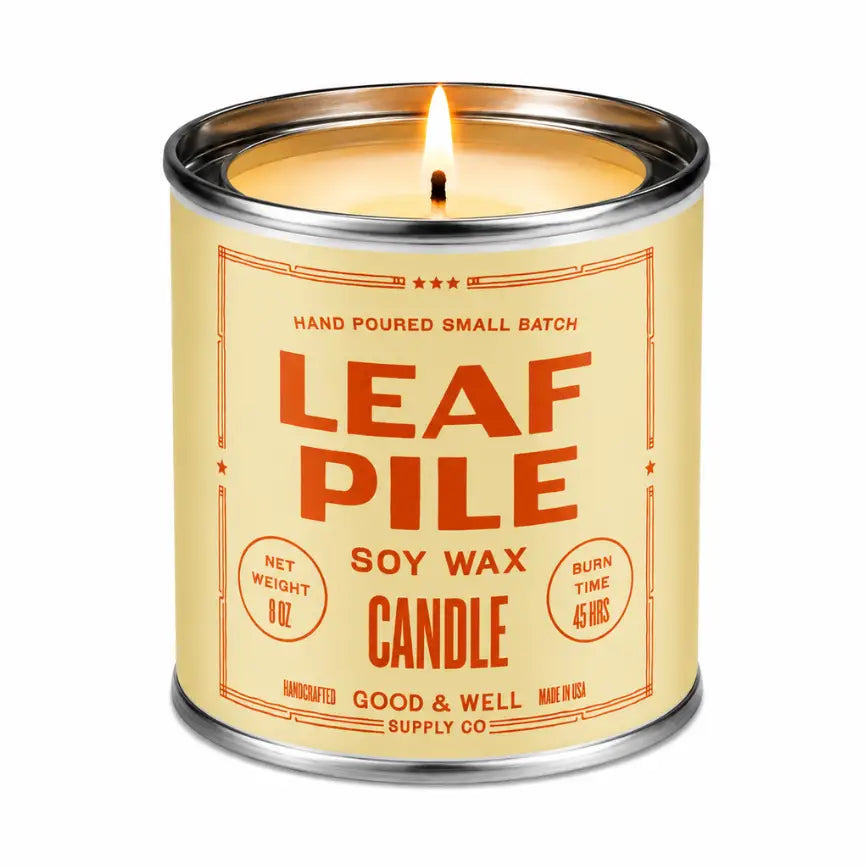 A lit soy wax candle in a cylindrical metal tin with a cream-colored label and orange-red text reading 'LEAF PILE' and 'SOY WAX CANDLE' on a white background.