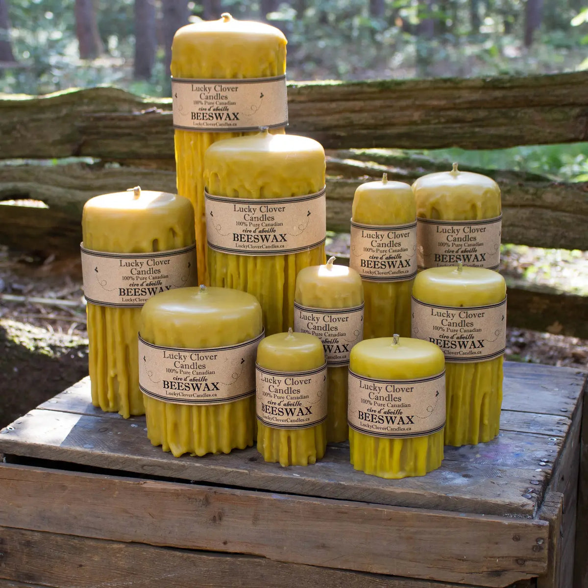 A group of yellow beeswax pillar candles in various sizes, arranged on a rustic wooden crate outdoors with a blurred forest background.