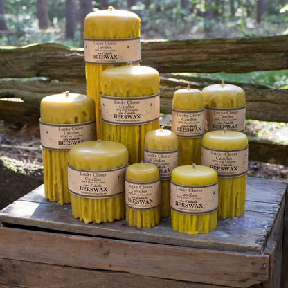 A group of yellow beeswax pillar candles in various sizes, arranged on a rustic wooden crate outdoors with a blurred forest background.