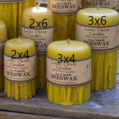 Several yellow beeswax pillar candles with kraft paper labels are arranged on a rustic wooden surface. The labels show the brand name Lucky Clover Candles and sizes including 2x6, 3x6, and 3x4.