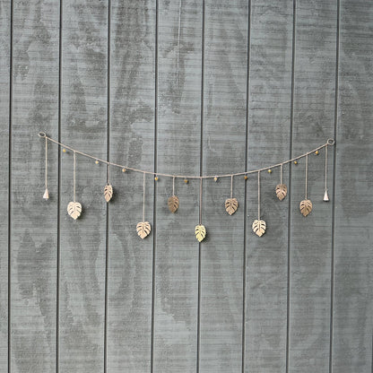 A decorative string garland with hanging metallic leaf ornaments, displayed against a gray wooden plank wall.