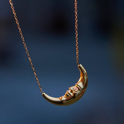 A gold crescent moon pendant necklace hanging against a dark blue blurred background.