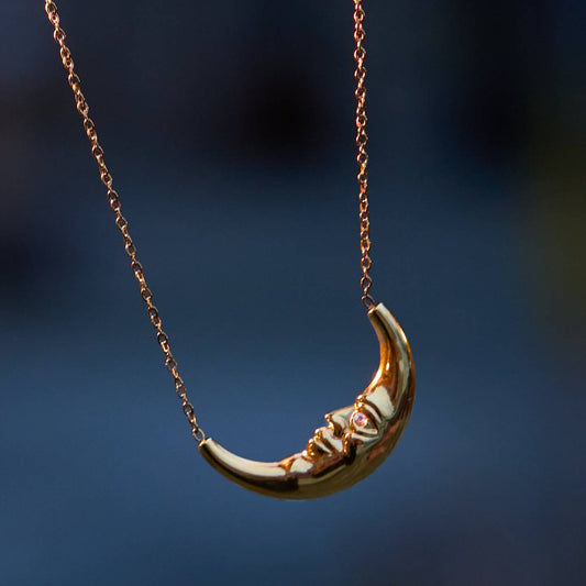 A gold crescent moon pendant necklace hanging against a dark blue blurred background.