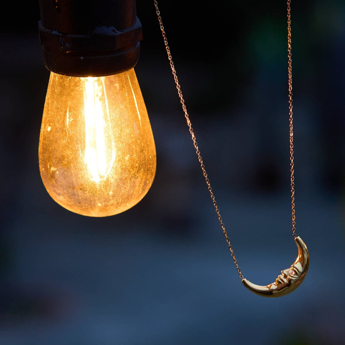 A gold crescent moon pendant necklace hanging next to a lightbulb, against a dark blue blurred background.